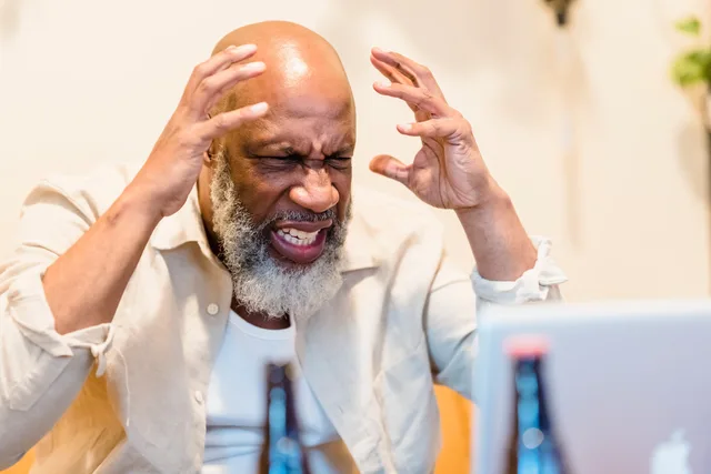 An adult man expressing frustration while sitting in front of a laptop in an indoor setting. Eine konkrete Beschwerde statt nur den Ton beantworten