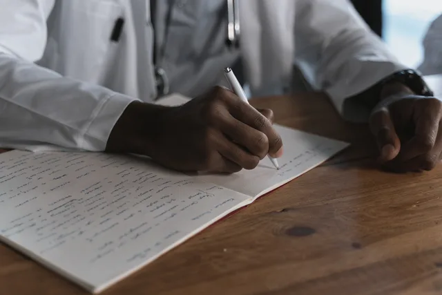 Close-up of a doctor writing notes with a pen in a medical notebook on a wooden desk. Entscheidungen während eines Vorfalls dokumentieren