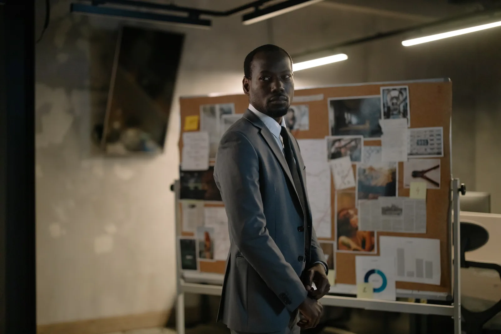 A serious man in a suit stands in an office, examining a bulletin board with notes and photos.
